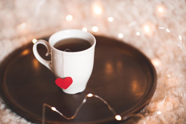 Taza de té con corazón rojo en una bandeja de madera sobre luces en el fondo Día de San Valentín Buenos días