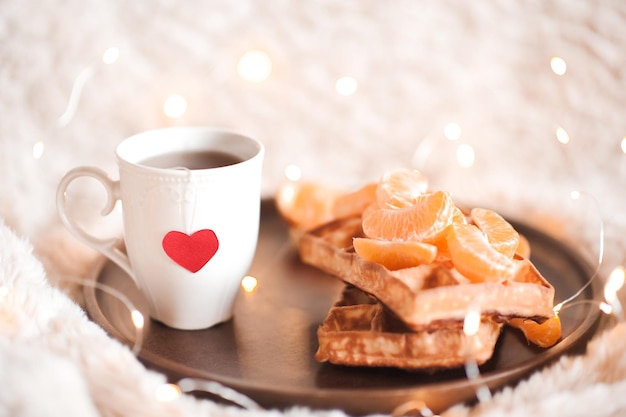 Taza de té con corazón rojo con gofres y naranjas en bandeja de madera en la cama Desayuno sabroso Buenos días