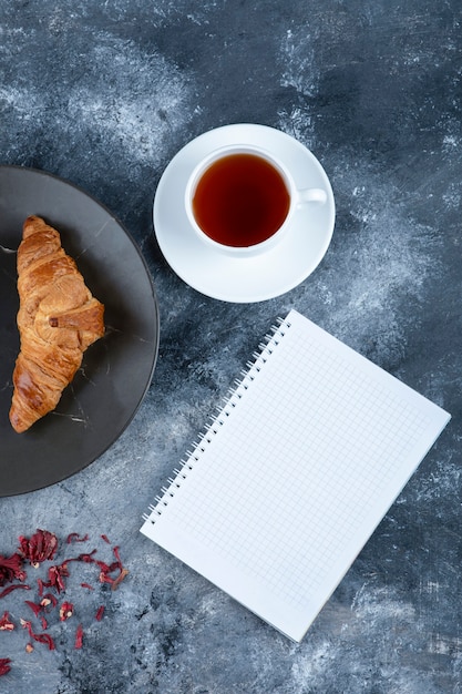 Una taza de té con croissant y cuaderno abierto colocado sobre la mesa de piedra.