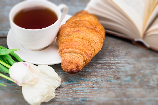 Taza de té con croissant, tulipanes frescos y libro sobre un fondo de madera. Imagen perfecta para el día de la madre.