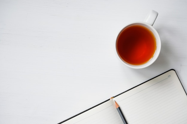 Taza de té con el cuaderno y el lápiz en la tabla de madera blanca.