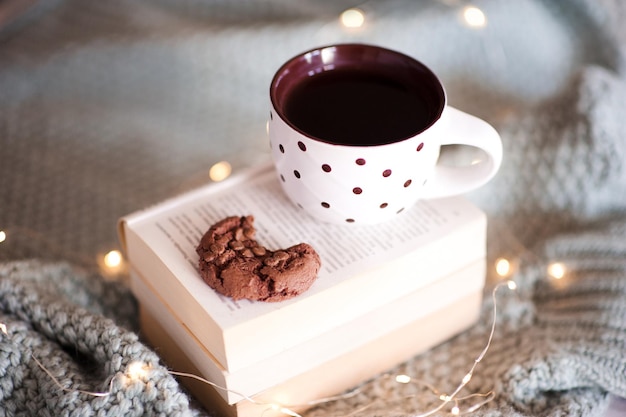 Taza de té con galleta de chocolate con mordedura de libros abiertos sobre las luces de Navidad de cerca. Temporada de vacaciones de invierno. Buenos días. Año nuevo.