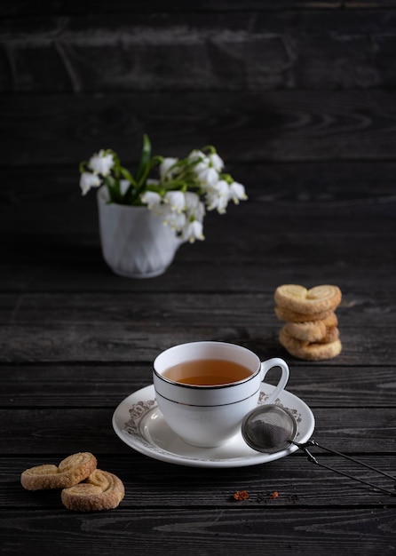 Una taza de té con galletas y un colador de té sobre un fondo oscuro con racimos de flores blancas de gotas de nieve