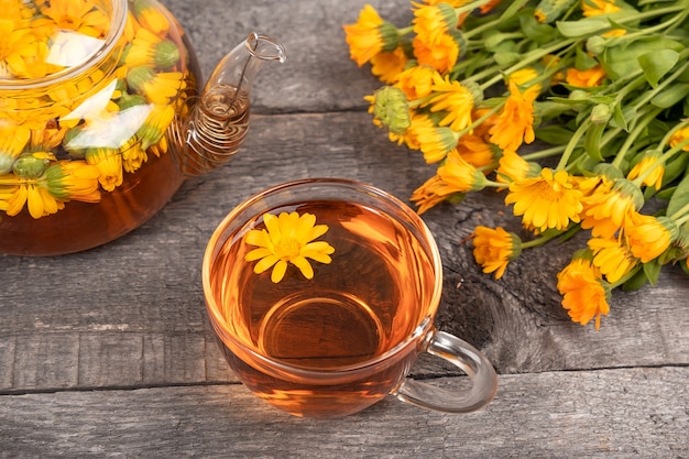 Taza de té de hierbas y tetera transparente y flores de caléndula sobre fondo de madera. El té de caléndula beneficia su concepto de salud.