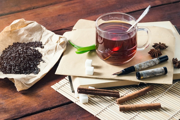 Taza con té, hojas de té secas sobre papel artesanal, sobre de rebanadas de azúcar y manija sobre una mesa de madera