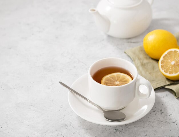 Una taza de té con limón y una cuchara sobre un fondo claro con una tetera blanca y cítricos Concepto de bebida saludable por la mañana Espacio libre para texto