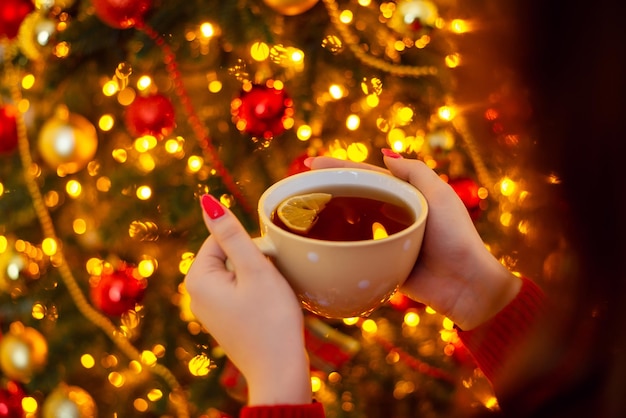 Una taza de té con limón en manos de una joven hermosa y cálidas luces de árbol de navidad en el fondo Año nuevo navidad acogedor ambiente mágico festivo en casa