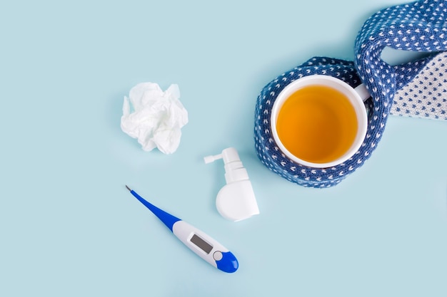 Taza de té de manzanilla, termómetro, servilleta arrugada y spray para secreción nasal y garganta sobre fondo azul. Enfermedades estacionales y tratamiento de resfriados, gripe, calor. Prevención de virus, copie el espacio para el texto.