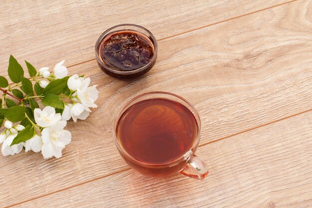 Taza de té, mermelada de fresa casera en un recipiente y flores de jazmín blanco sobre fondo de madera. Vista superior con copia espaciada.