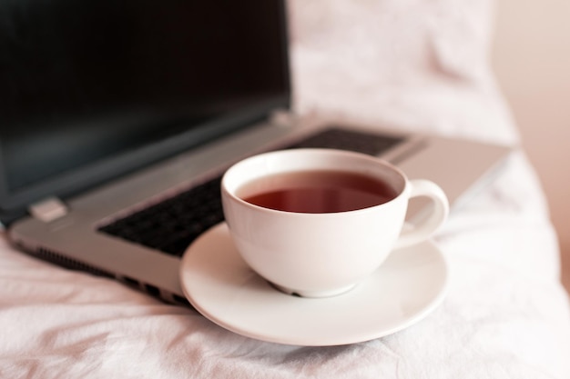 Taza de té negro en la computadora en primer plano de la cama. Buenos días. Hora del desayuno. Distanciamiento social. Quédate en casa.