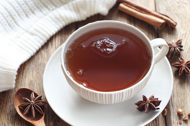 Una taza de té negro se encuentra sobre una mesa de madera. Cerca se encuentra un suéter blanco, anís estrellado y palitos de canela.