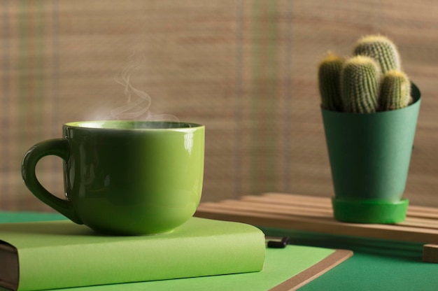 taza de té o café de color verde caliente con libro de vapor y cactus con mesa