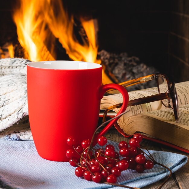 Una taza de té o café, una cosa de lana y bayas de viburnum cerca de una acogedora chimenea, en una casa de campo, en las vacaciones de otoño o invierno.