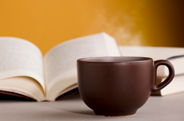 Taza de té o café marrón humeante caliente en el escritorio con libro abierto y fondo amarillo anaranjado