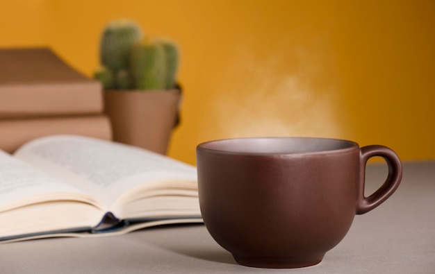 Taza de té o café marrón humeante caliente en el escritorio con libro abierto y fondo amarillo naranja de cactus