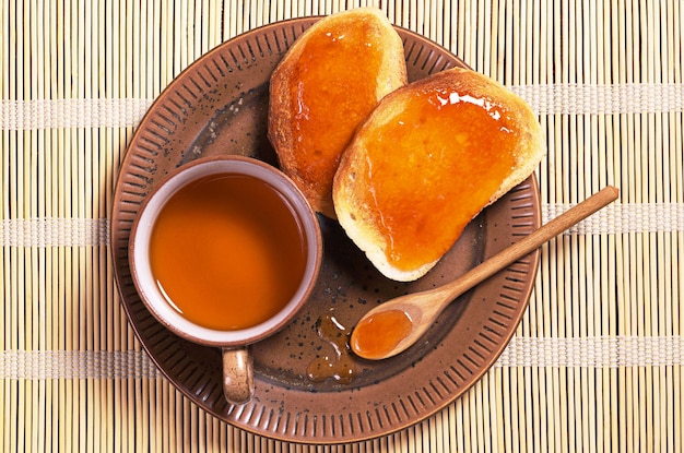 Taza de té y pan blanco asado con mermelada en plato sobre servilleta de bambú, vista superior