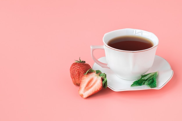 Taza de té en un plato blanco con hojas de menta y fresas maduras. fondo rosa copyspace