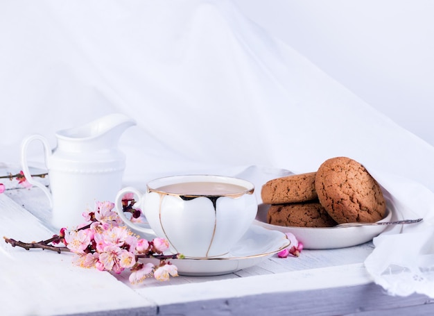 Taza de té de porcelana blanca jarra de leche y galletas de avena recién horneadas Desayuno inglés bodegón con bebida y golosinas y mantel