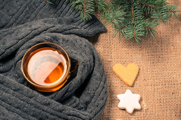 Taza de té con rodaja de limón envuelta en bufanda de lana, galletas de jengibre en forma de estrella y corazón, ramas de abeto natural y canela, anís estrellado sobre tablas de madera. Efecto tonificante de color.