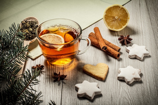 Taza de té con rodaja de limón, galletas de jengibre en forma de estrella y corazón, ramas de abeto natural con cono y canela, anís estrellado sobre tablas de madera. Efecto tonificante de color.