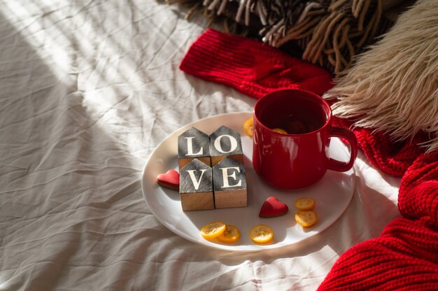Taza de té rojo con kumquat en el plato y galletas de dos corazones con la palabra AMOR en una cama blanca