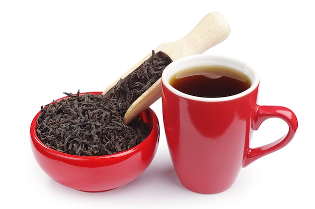 Taza de té y té negro seco en un recipiente rojo con una cuchara de madera sobre blanco