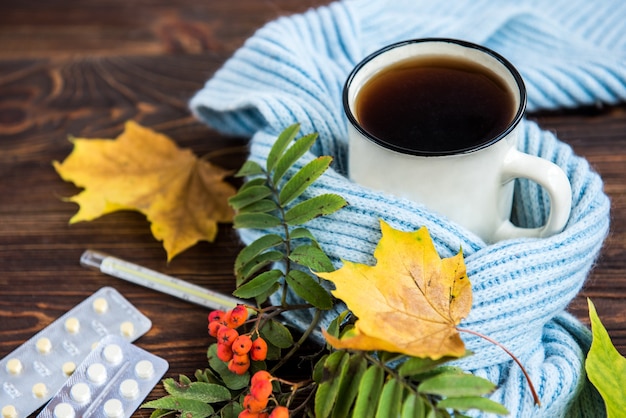 Taza de té con termómetro, bufanda azul y hojas de otoño sobre fondo de madera. Temporada de gripe en otoño, enfermedad.