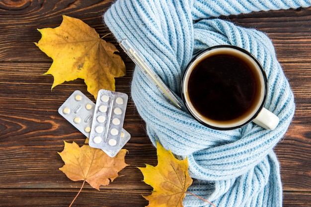 Taza de té con termómetro, bufanda azul y hojas de otoño sobre fondo de madera. Temporada de gripe en otoño, enfermedad.