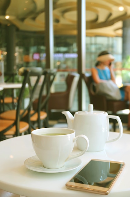 Taza de té verde caliente y tetera blanca con un teléfono móvil en la mesa del café