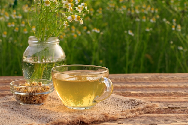 Taza de té verde, tarro con flores de manzanilla blanca y cuenco de cristal con flores secas de matricaria chamomilla sobre tela de saco y tablas de madera con fondo verde natural.