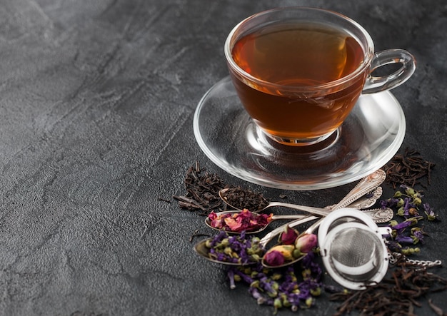 Taza de té de vidrio con infusor y varios tés sobre fondo negro Brotes de rosa de té sueltos negros y verdes Flores de malva azul