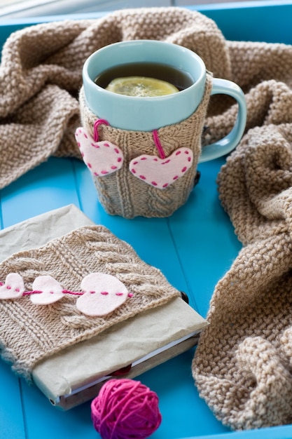 Una taza de té en un viejo cuaderno de suéter con corazones de manta de punto beige de fieltro y radios sobre fondo azul