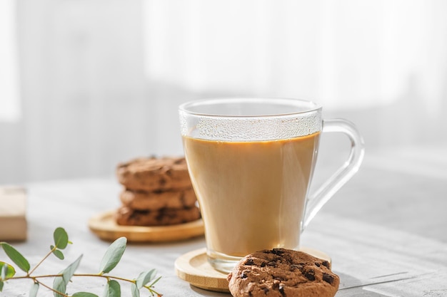 Una taza transparente de café con leche con galletas de chocolate y hojas de eucalipto en las sombras matutinas y los rayos del sol a través de la ventana en el fondo gris de la mesa Concepto de café matutino de cerca