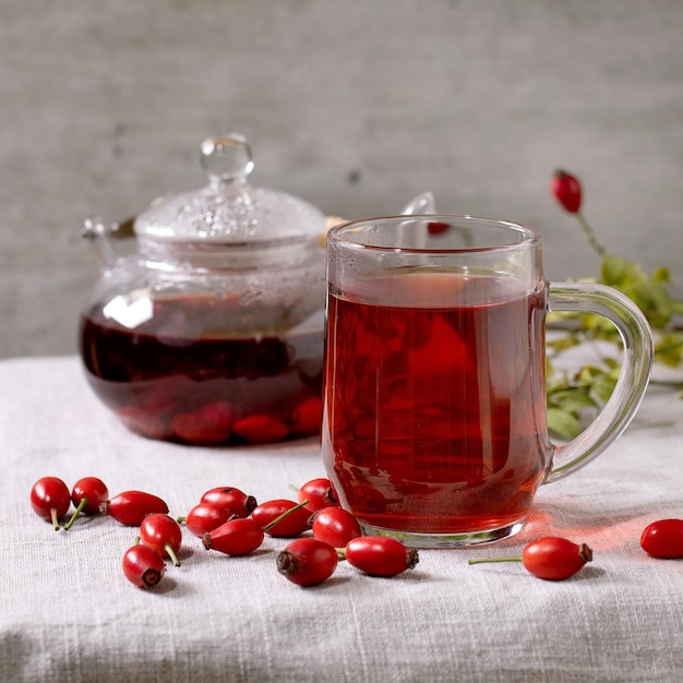 Taza transparente de té de hierbas de bayas de rosa mosqueta y tetera de vidrio de pie sobre un mantel de lino blanco con bayas silvestres de otoño alrededor de la bebida cálida y acogedora de invierno Imagen cuadrada