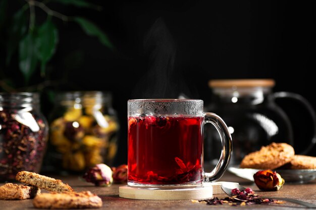 Taza transparente con té rojo caliente en un oscuro, cerca de galletas y hojas de té