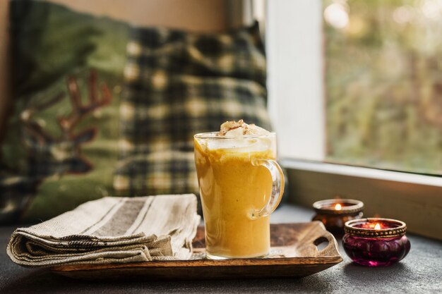 Una taza de vidrio de calabaza con leche junto a una ventana