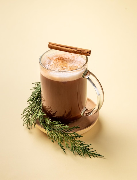 Taza de vidrio de chocolate caliente con crema batida y bastón de canela en un soporte con ramas de pino en un fondo amarillo