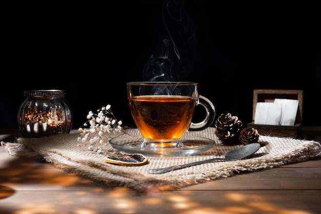 Taza de vidrio con té caliente sobre una mesa de madera con flores secas de limón y velas sobre fondo negro