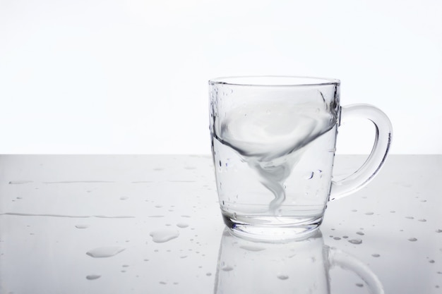 Taza de vidrio transparente con agua sobre una superficie clara con gotas de agua. Agua en forma de tornado dentro de la taza, enfoque suave. Una tormenta en una taza de té