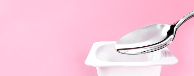 Taza de yogur y cuchara de plata sobre fondo rosa, recipiente de plástico blanco con crema de yogur, productos lácteos frescos para una dieta saludable y un equilibrio nutricional