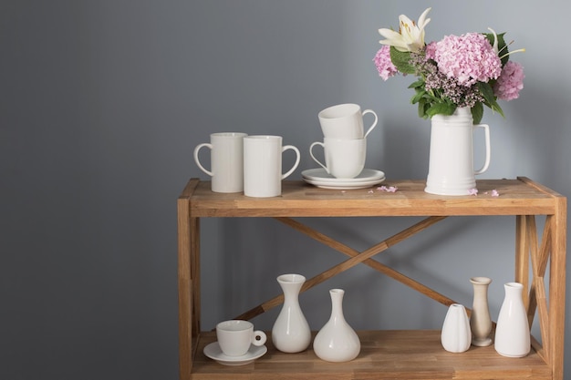 Tazas blancas y flores en un jarrón sobre un estante de madera sobre fondo gris.