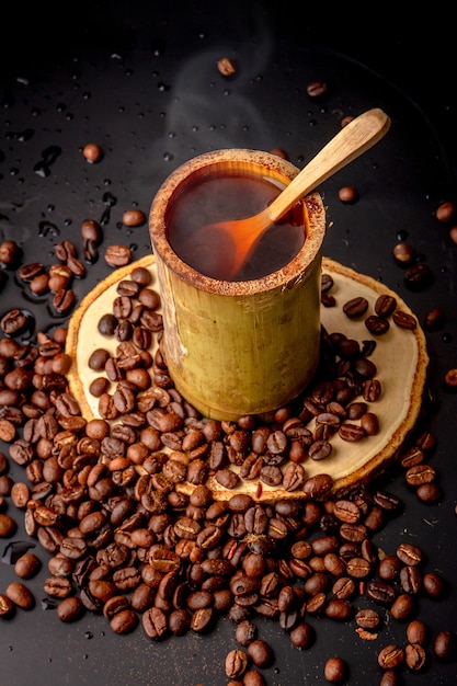 Tazas de café hechas de bambú en platillo de madera y muchos granos de café tostados esparcidos sobre fondo negro, el café oscuro tiene humo y aroma, bebida para el descanso, la mañana y la hora del café, estilo local.