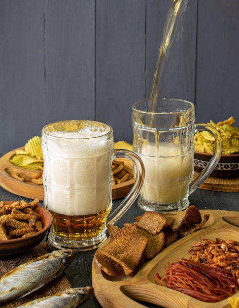 Tazas de cerveza fría con espuma servidas en una mesa de madera con bocadillos salados en un tazón de madera