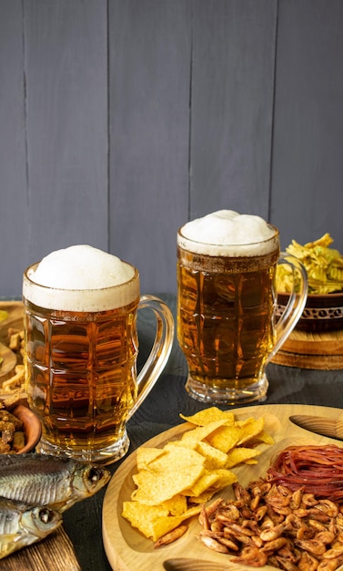 Tazas de cerveza fría con espuma servidas en una mesa de madera con bocadillos salados en un tazón de madera