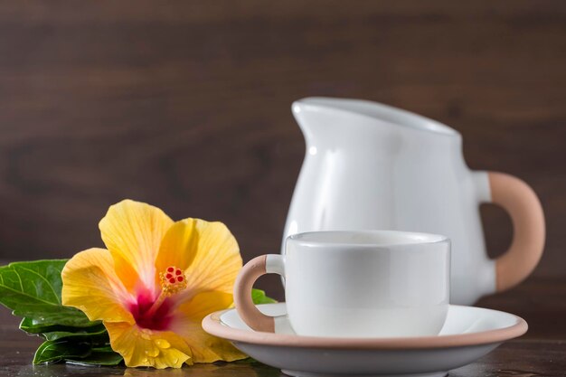 Tazas y jarra de leche junto a una flor de hibisco