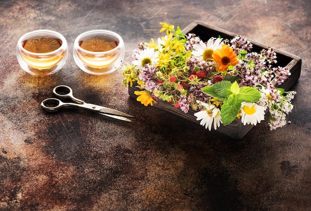 Tazas de té de hierbas y hierbas curativas en una bandeja de madera y tijeras sobre un fondo de hormigón marrón