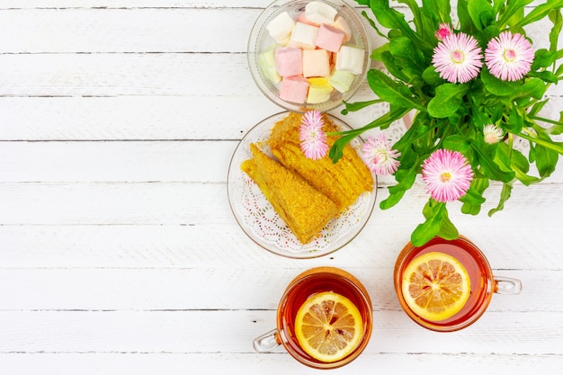 tazas de té, pedazos de pastel, ramo de flores rosadas y malvaviscos en una mesa de madera blanca