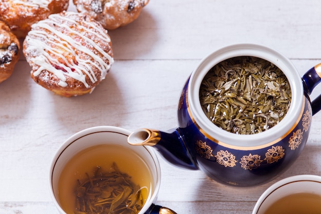 Tazas de té con té preparado, tetera y muffins en mesa de madera blanca