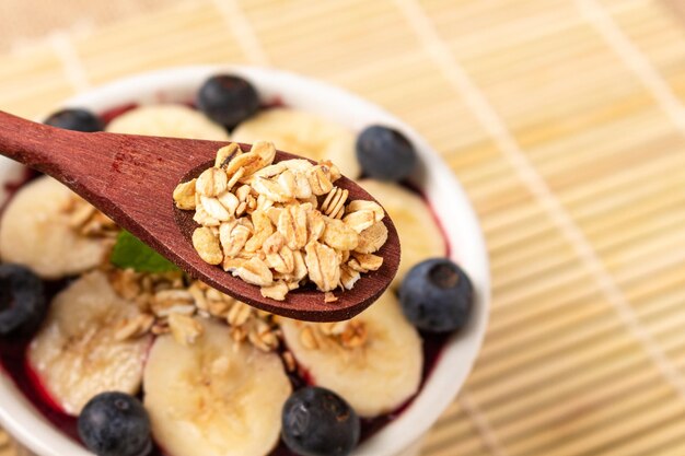 Tazón de Acai con plátano, arándano y cucharada de avena en la superficie de bambú.