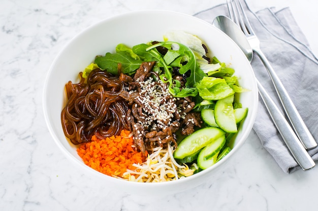 Tazón de almuerzo asiático con carne de res. fideos, mezclar hojas verdes y vegetales en la mesa de mármol. De cerca